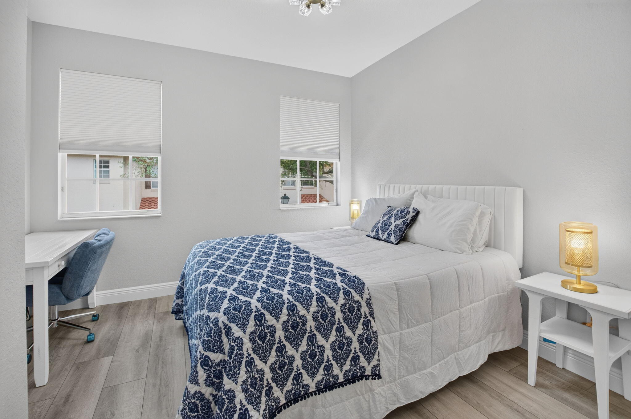 Bedroom with a white bed, blue patterned quilt, two windows, and a desk with blue chair.