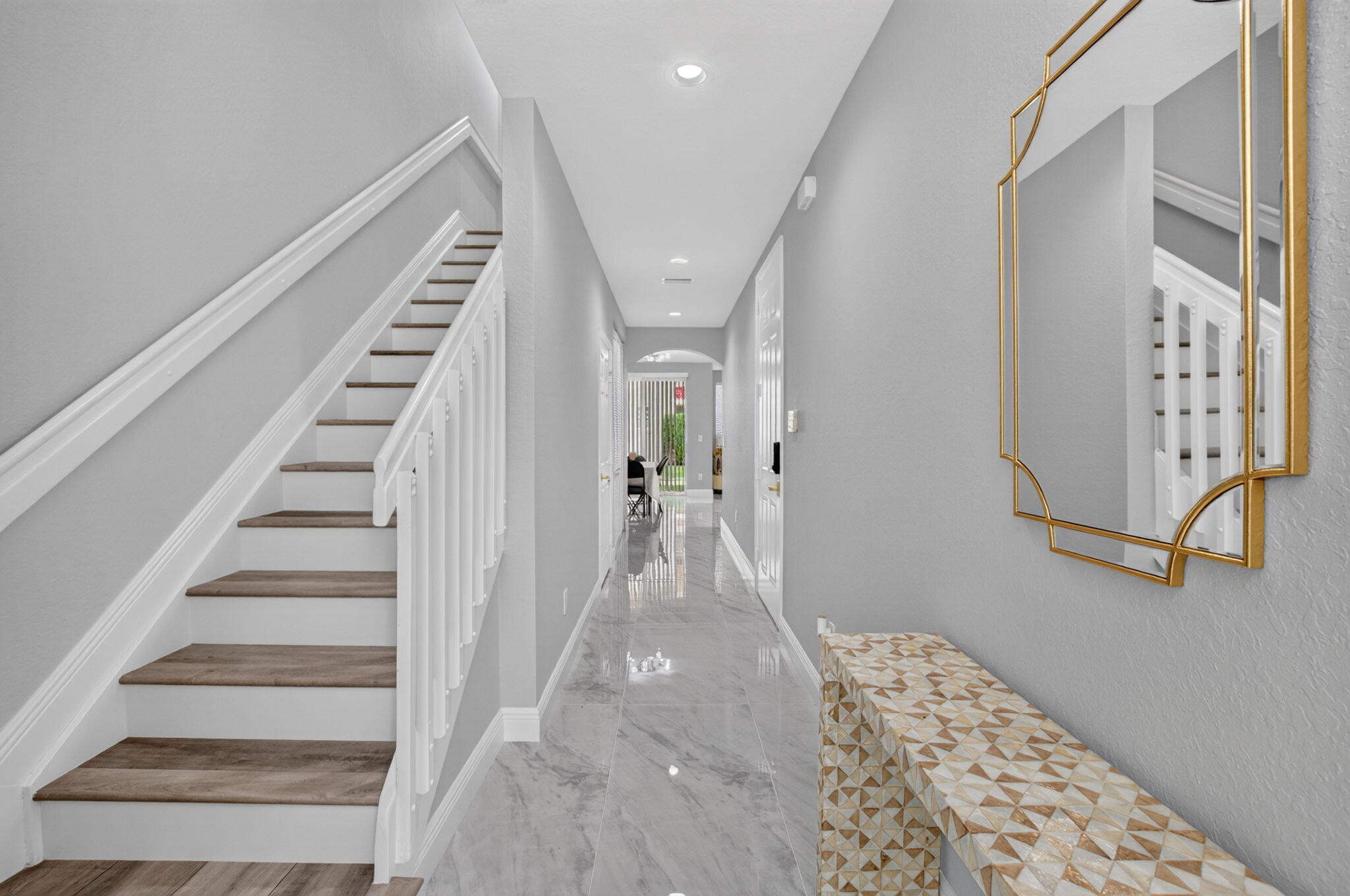 Entry hall with a staircase, polished gray tile, and a geometric console table beneath a gold-framed mirror.