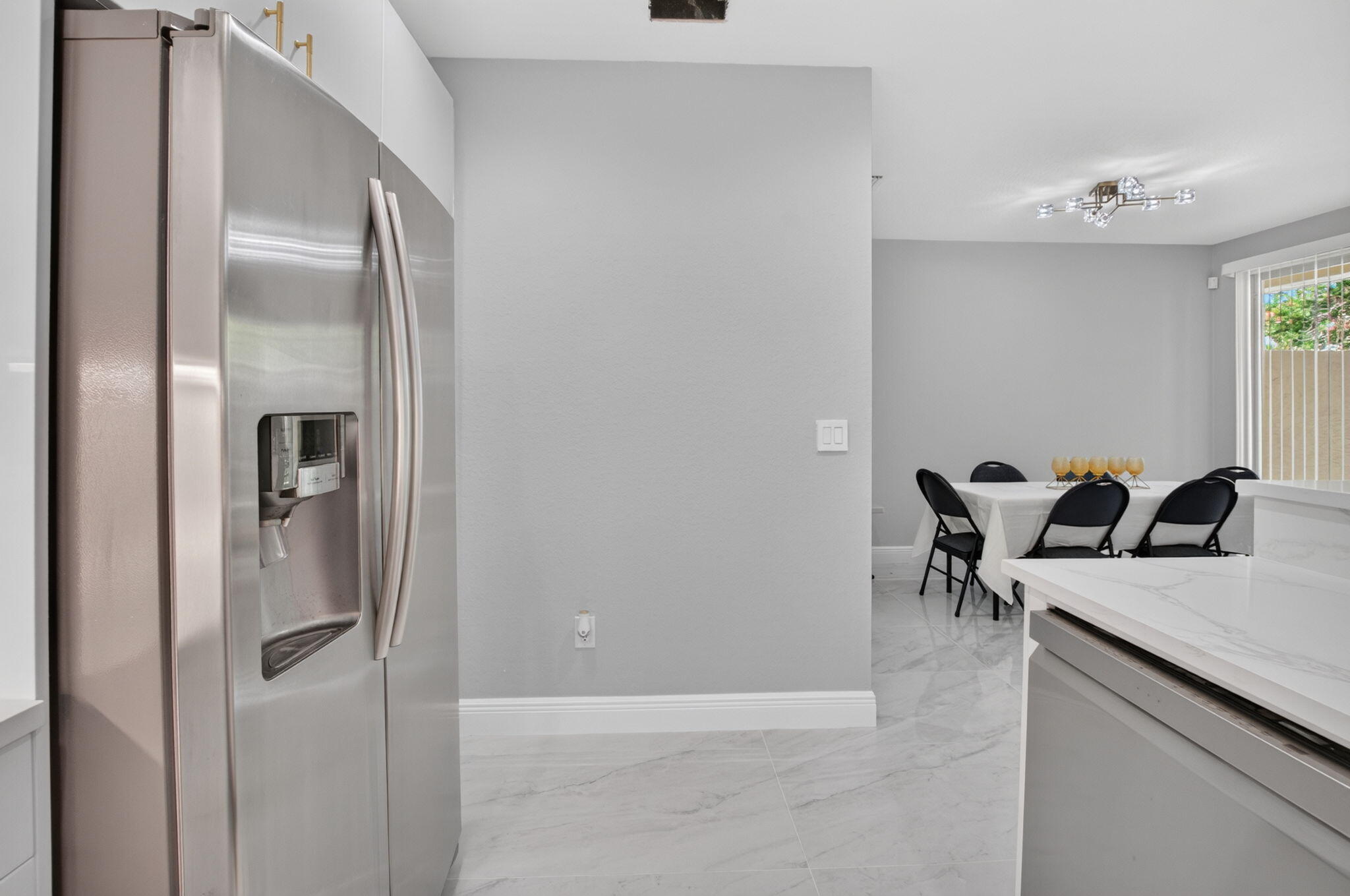 Modern kitchen with stainless steel refrigerator, marble-look island, and dining table by a window.