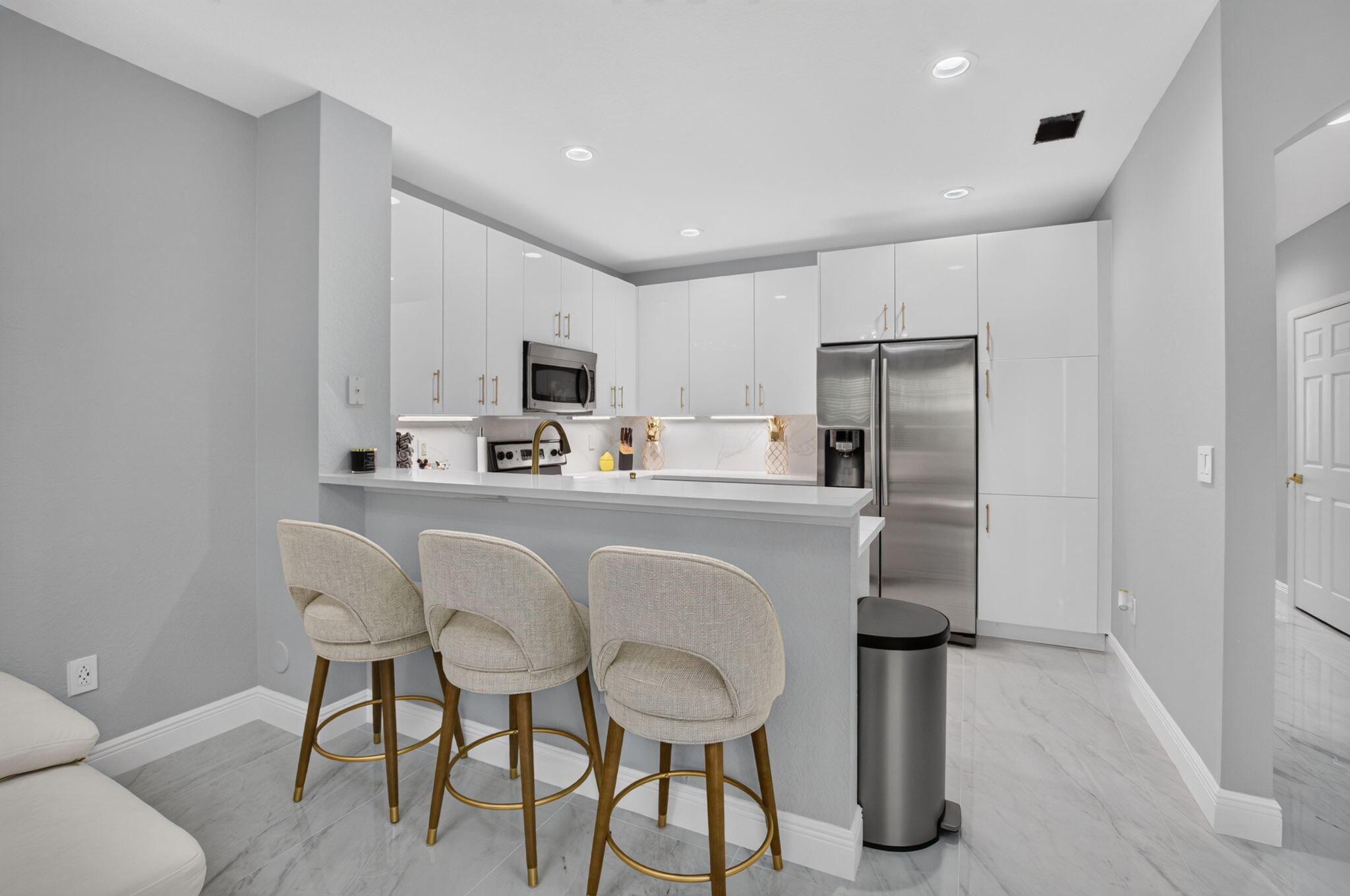 Modern white kitchen with stainless steel appliances and a breakfast counter lined with three upholstered stools.