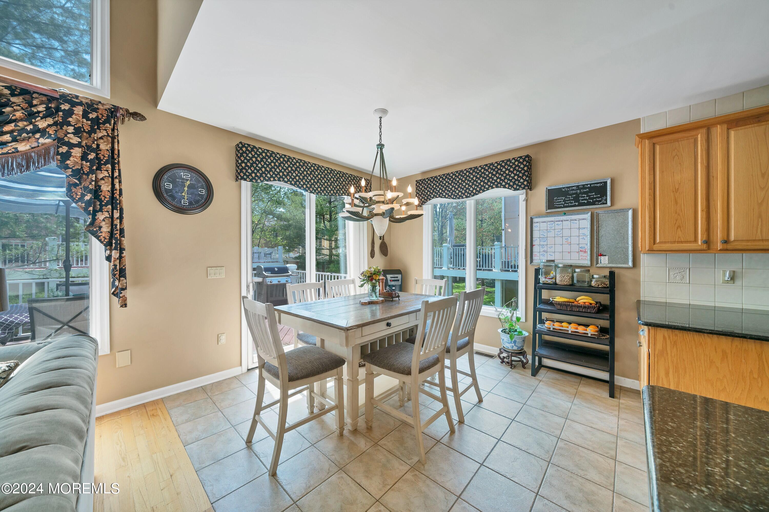 Dining area with a large table beneath a chandelier, beside windows overlooking a deck and wooded yard.