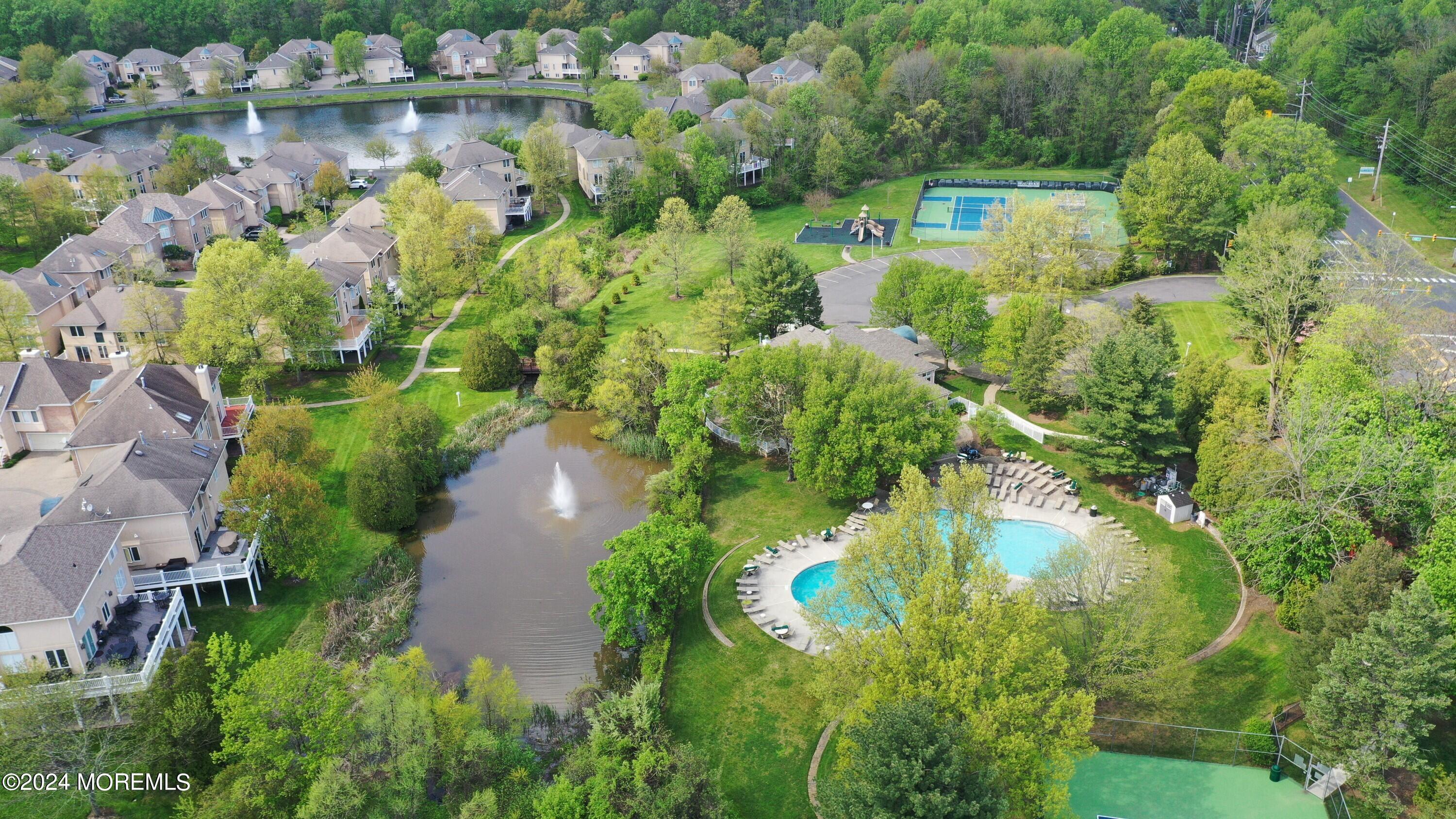 Aerial view of homes around a pond, pool, tennis courts, playground, and landscaped walking paths.