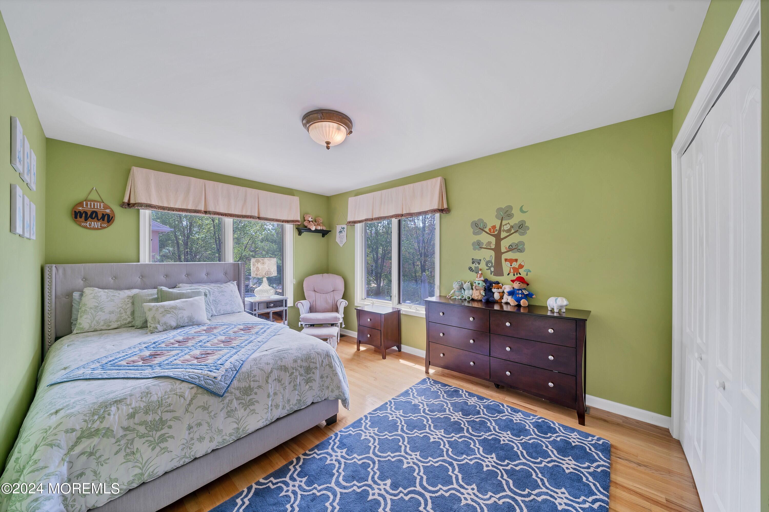 Bedroom with green walls, a gray bed, blue patterned rug, and windows overlooking trees.