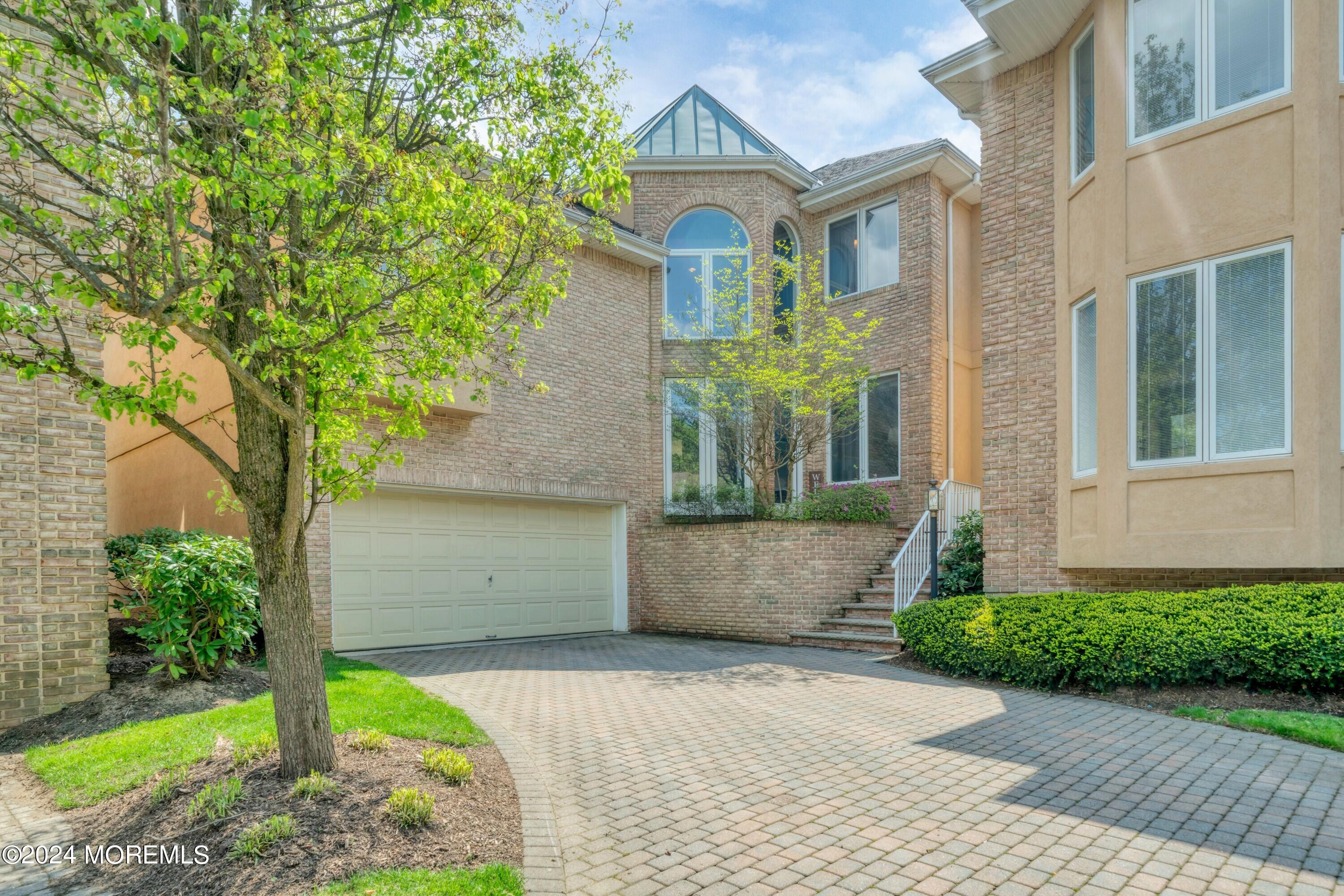 Two-story brick home with arched windows, double garage, and wide paver driveway framed by spring greenery.