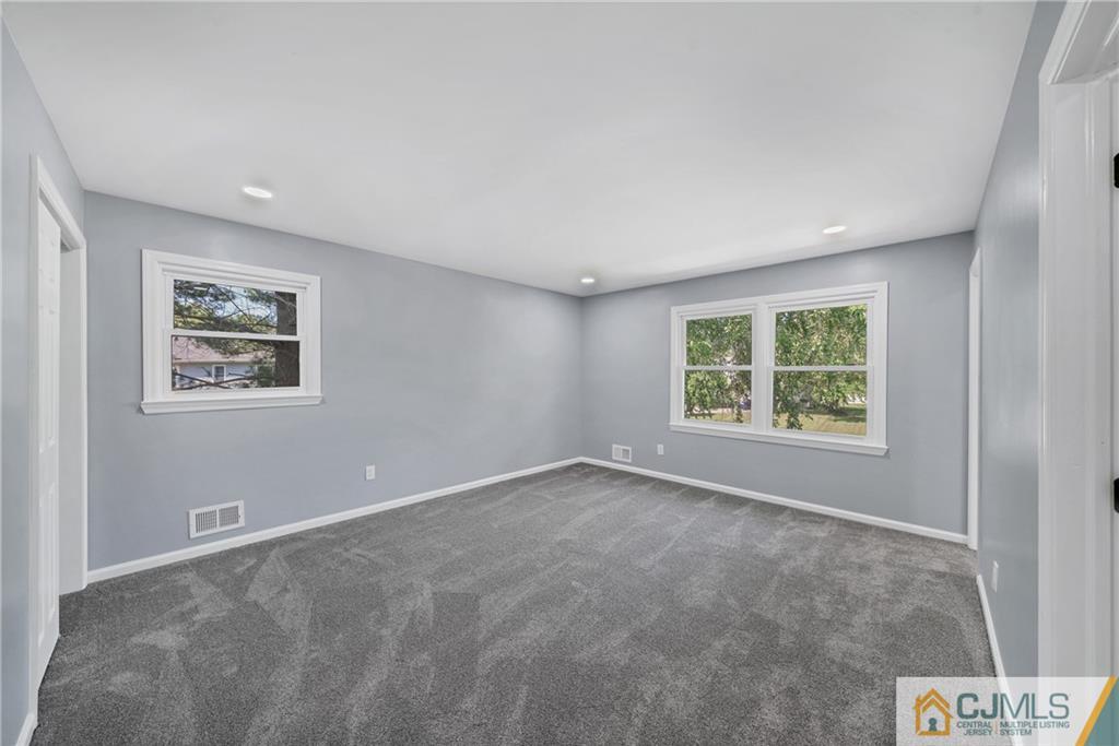 Empty bedroom with gray carpet, recessed lights, and windows overlooking greenery.