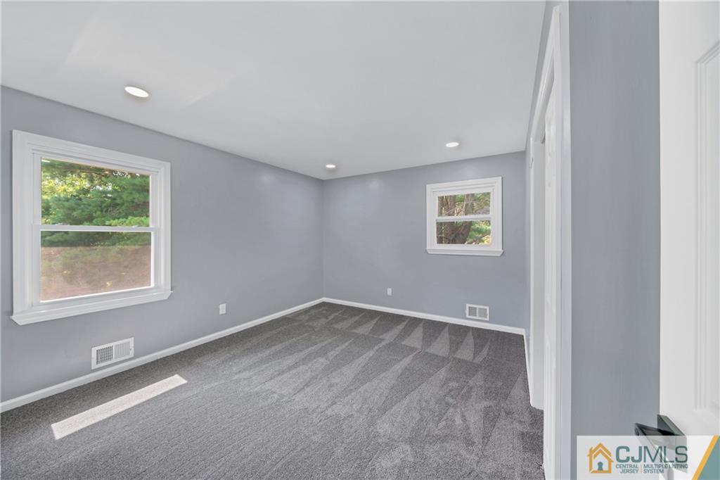 Empty bedroom with gray carpet, recessed lights, and two windows overlooking greenery.