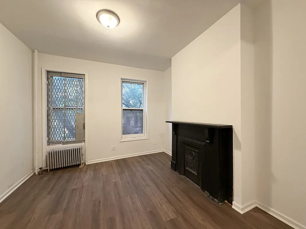 Vacant room with a black fireplace, dark hardwood floors, two windows and a radiator.