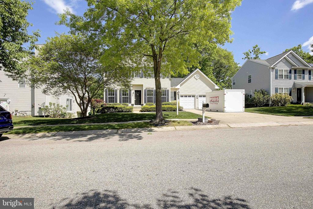 Two-story house with an attached garage, mature trees, and a PODS container in the driveway.
