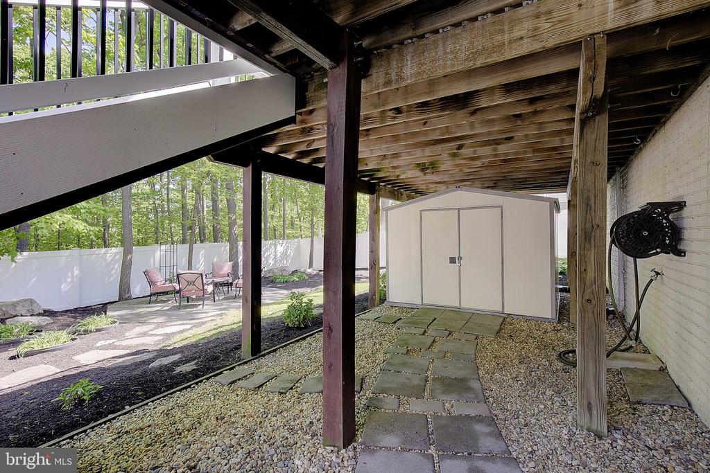 Covered backyard patio with a storage shed, stone path, and seating beside a white privacy fence.
