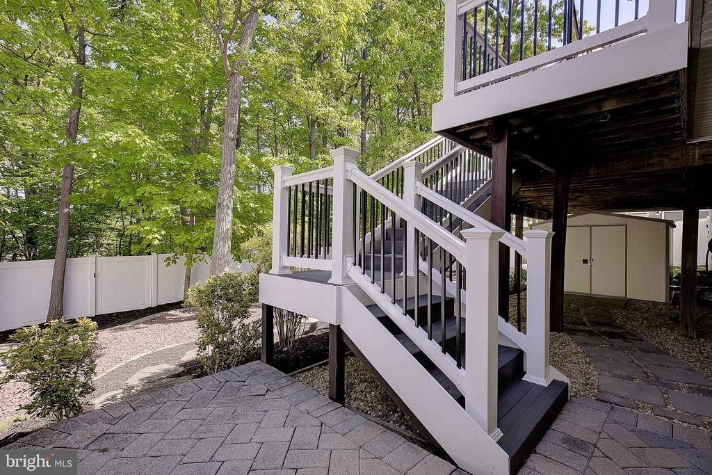 Raised deck with white-railed stairs above a paved patio in a leafy, fenced yard.