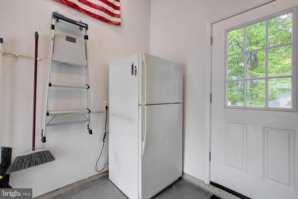 Garage with a white refrigerator, folding ladder, and windowed door overlooking leafy trees.