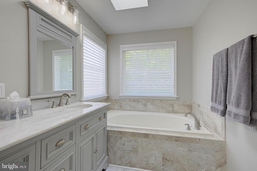 Bathroom with a tiled soaking tub, gray vanity, marble countertop, and skylight.