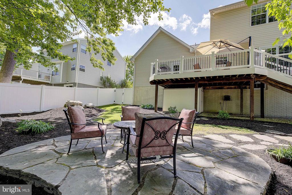 Backyard patio with cushioned chairs beneath a raised deck and umbrella, bordered by garden beds and a white fence.