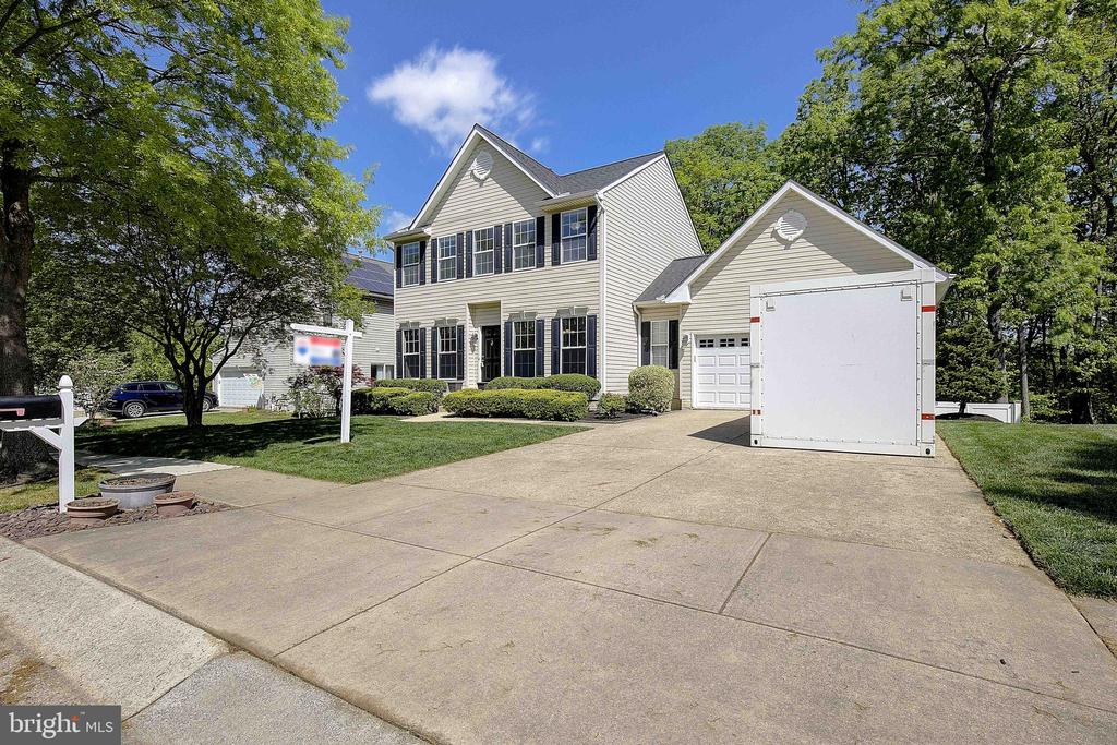 Two-story house with dark shutters, attached garage, and a broad driveway partly blocked by a white trailer.