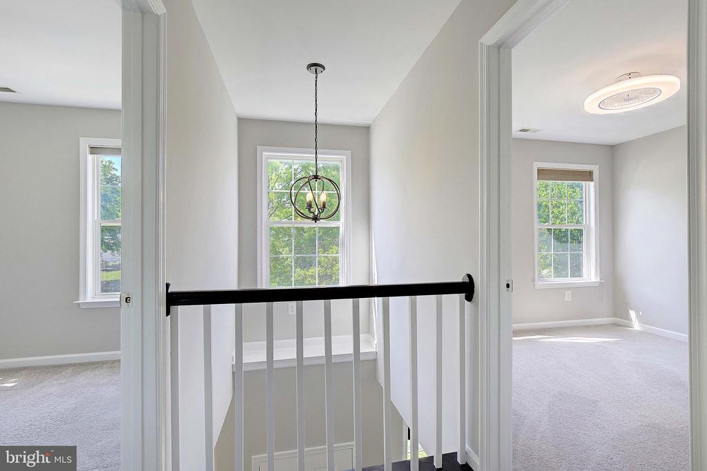 Upstairs landing with black railing, hanging chandelier, and bright carpeted rooms on either side.
