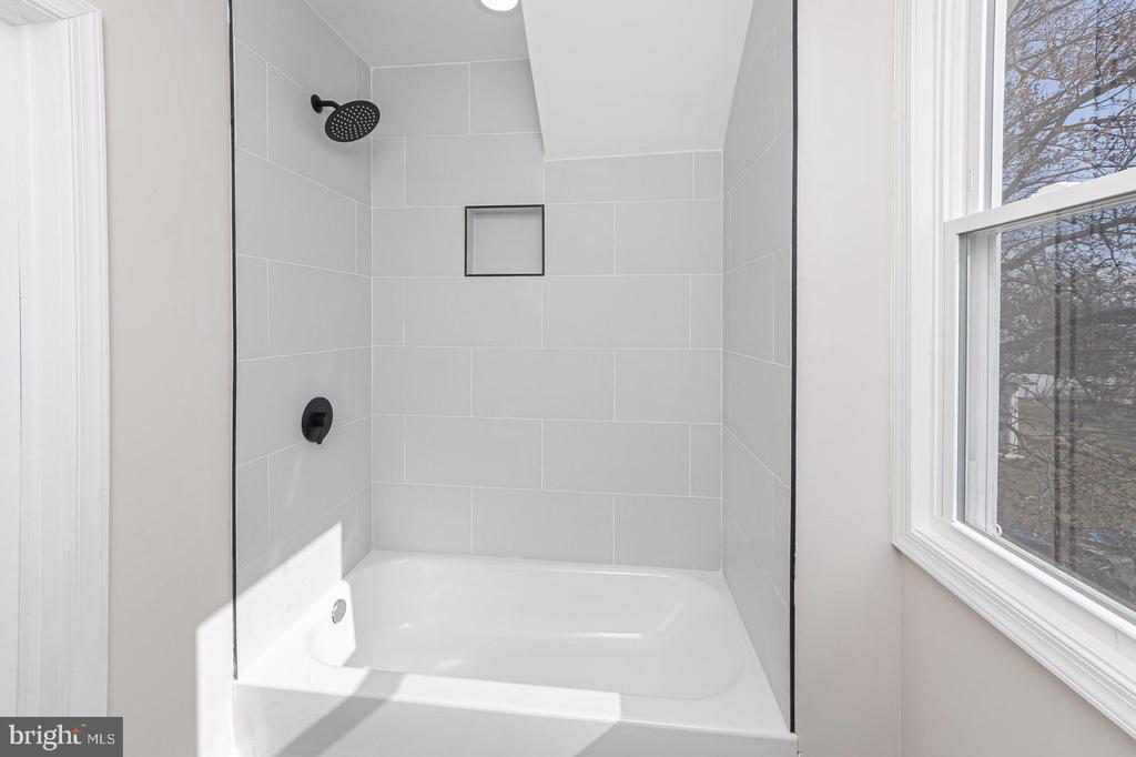 Bathroom shower with light-gray tile, matte-black fixtures, and a window beside the white tub.