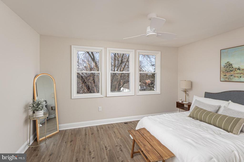Staged bedroom with a white bed, wood bench, and three windows overlooking trees.
