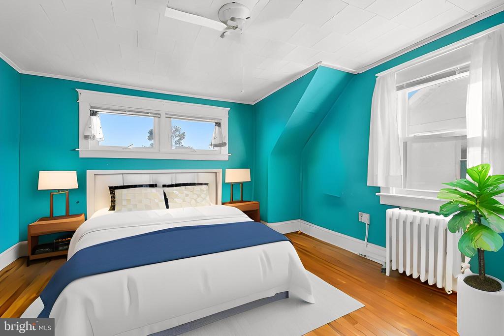 Staged bedroom with teal walls, a white bed with blue runner, hardwood floors, and curtained windows.