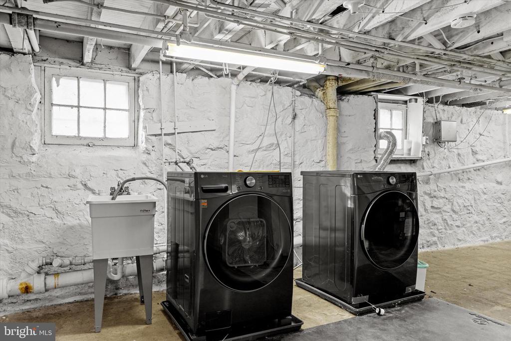 Basement laundry area with front-loading washer and dryer beside a utility sink and exposed stone walls.