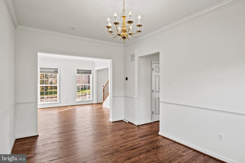 Empty dining room with hardwood floors and a brass chandelier, opening to a bright room with two windows.