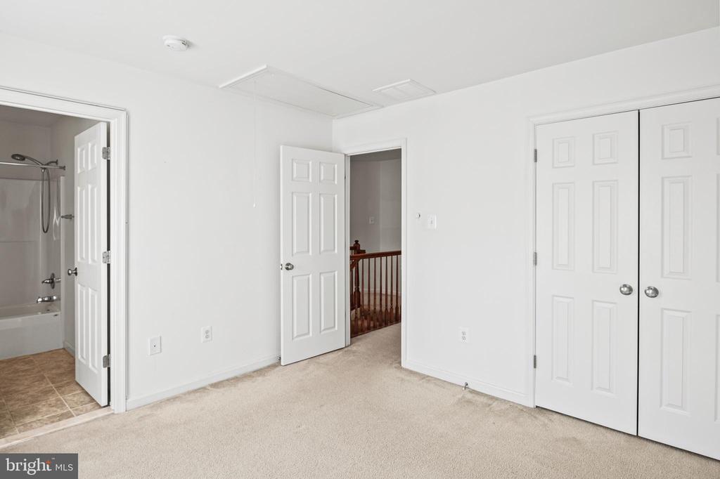 Carpeted bedroom with double closets, an open bathroom doorway, and a hallway railing.