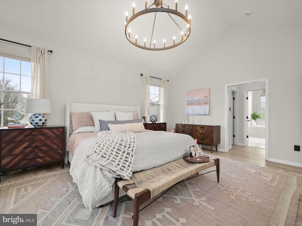 Staged bedroom with a white bed, patterned wood dressers, tall windows, and a circular chandelier.