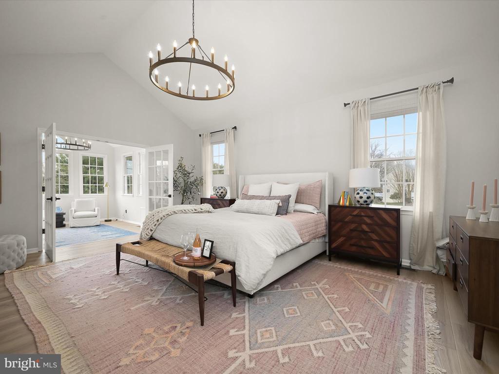 Staged bedroom with white and blush bedding, a brass ring chandelier, and French doors to a sitting room.