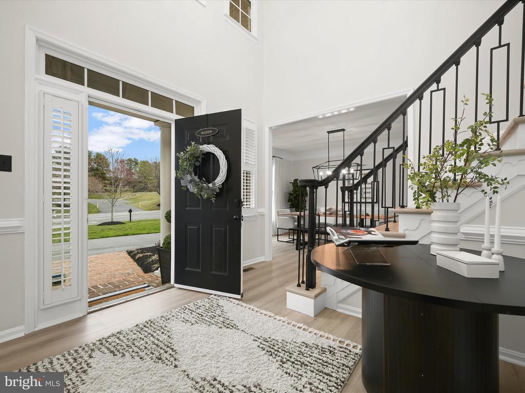 Entry foyer with open front door, patterned rug, and white staircase with black railings.