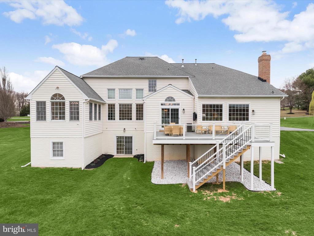 Rear of a white-sided home with a furnished raised deck, exterior stairs, and broad green lawn.