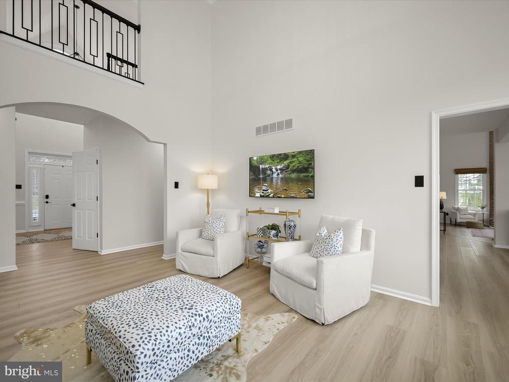 Staged living room with white armchairs, a patterned ottoman, and an arched entry beneath a loft railing.