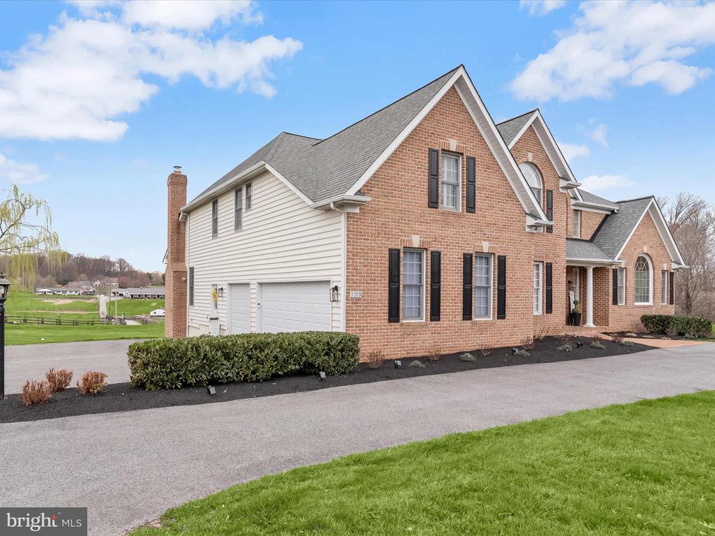 Two-story brick home with an attached garage, gabled roof, and paved driveway beside open fields.