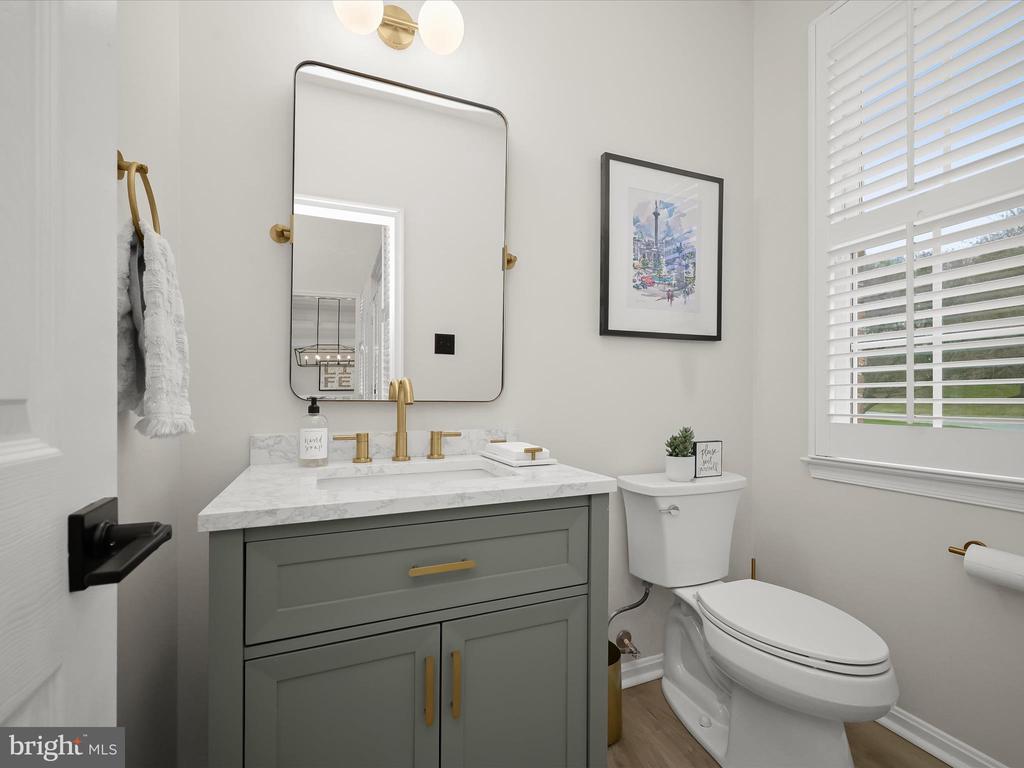 Bathroom with a gray vanity, marble countertop, brass fixtures, and a toilet beside a window with white blinds.