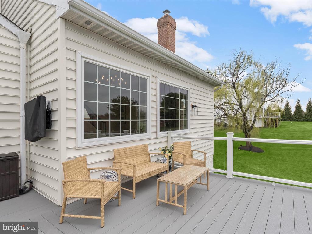 Furnished deck beside a white home, with wicker seating overlooking a green lawn and mature trees.