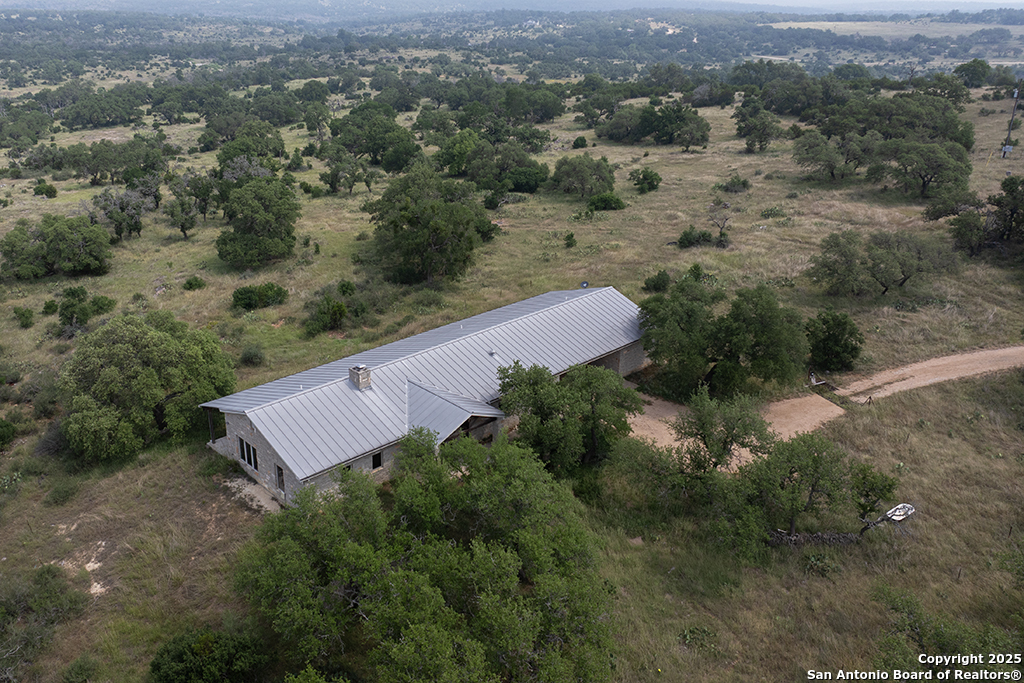 Aerial view of a rural stone house with a metal roof amid open, tree-dotted land.