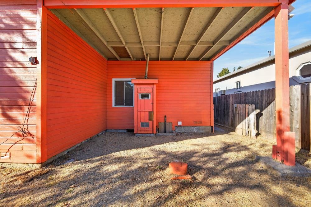 Covered backyard patio with red-orange siding, exposed roof beams, and a bare dirt yard.