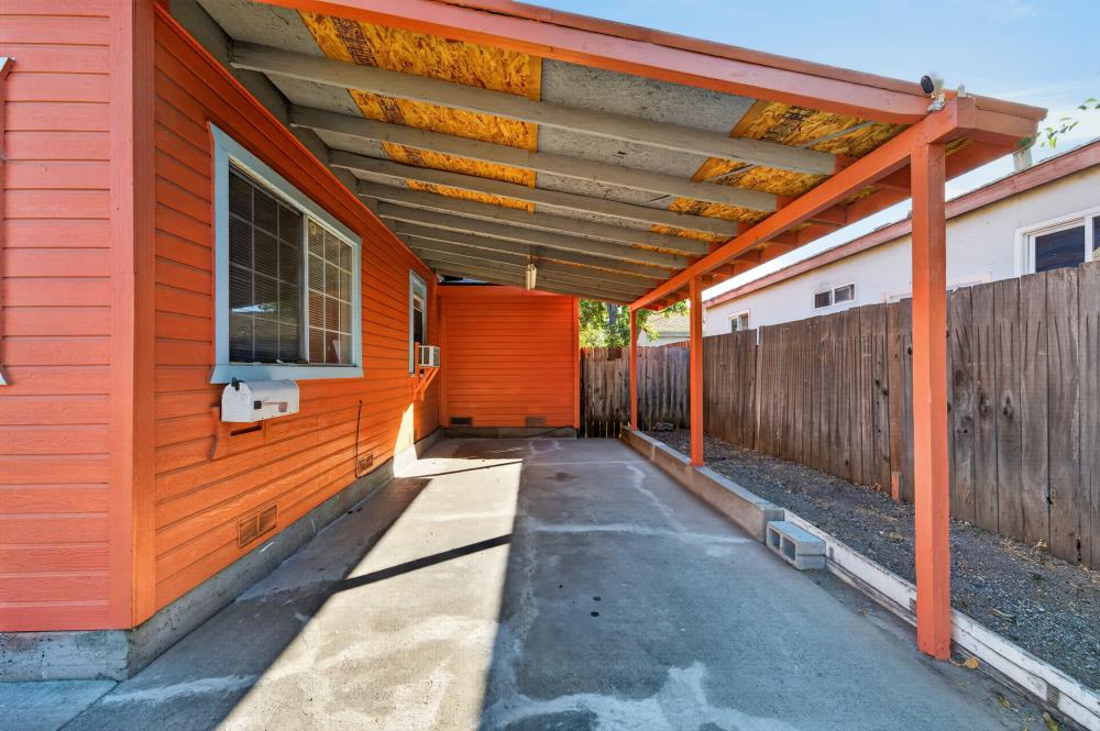 Covered carport beside a red-orange house, with exposed roof framing and a wooden privacy fence.