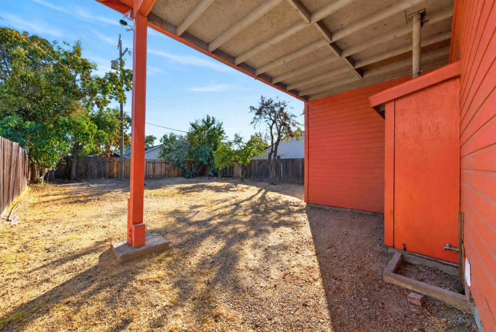 Dry fenced backyard beneath a covered patio beside a red-orange outbuilding, with mature trees.