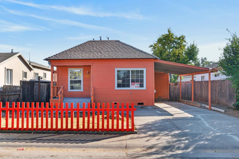 Red-orange single-story house with a gray shingle roof, covered carport, and bright red picket fence.