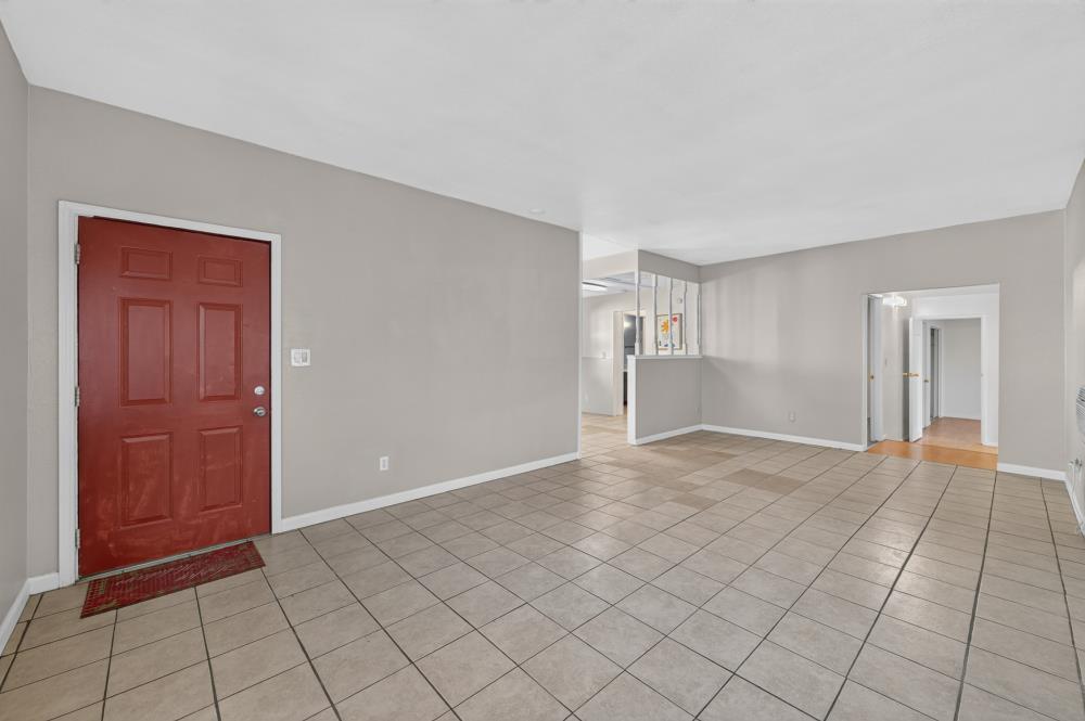 Empty tiled entry and living area with a red front door and openings to the kitchen and hallway.