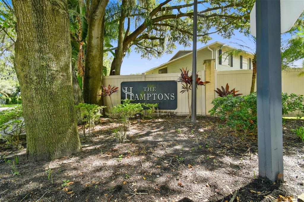 The Hamptons community entrance sign beneath mature oak trees, with a residential building behind it.