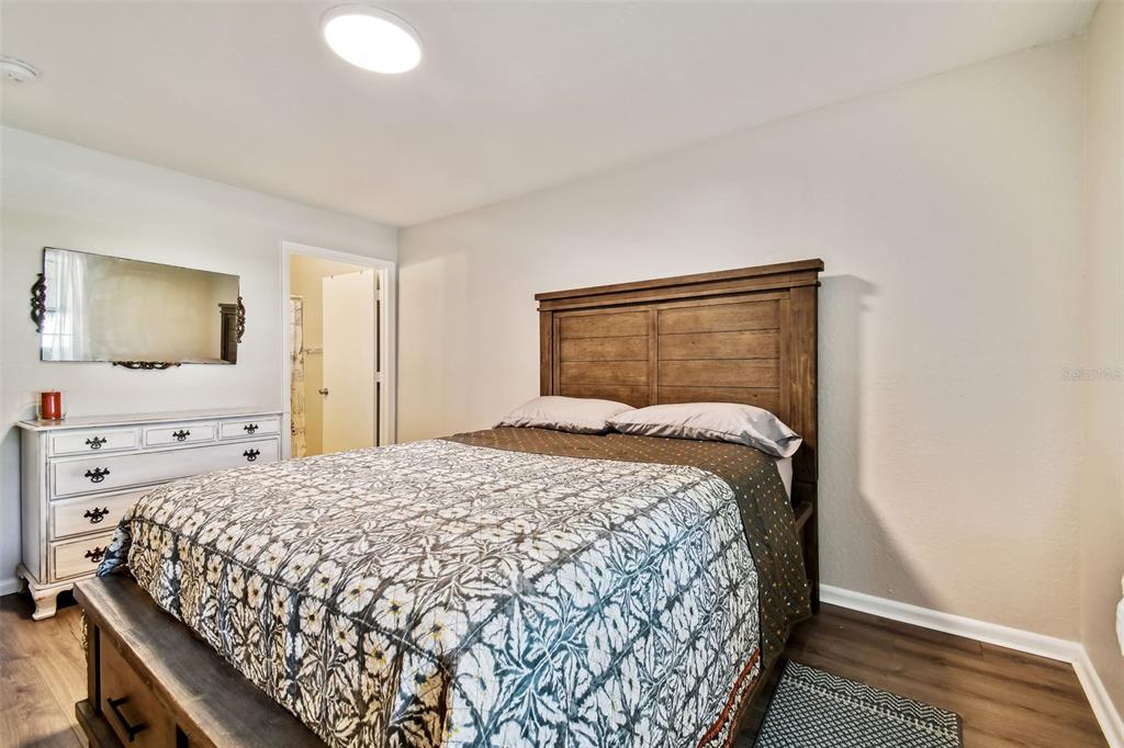 Furnished bedroom with a patterned bedspread, tall wooden headboard, and white dresser beneath a mirror.