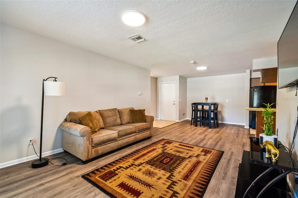 Living room with a tan sofa, patterned rug, and adjoining dining area and kitchen.
