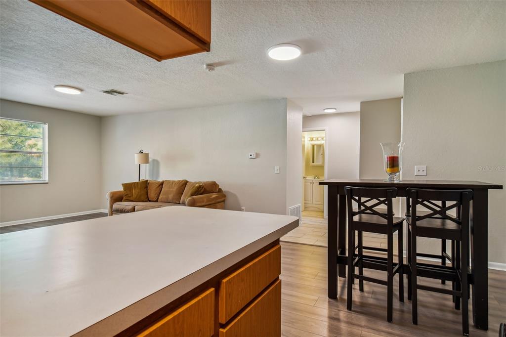 Open-plan living and dining area with a foreground kitchen island, brown sofa, and bar-height table with stools.