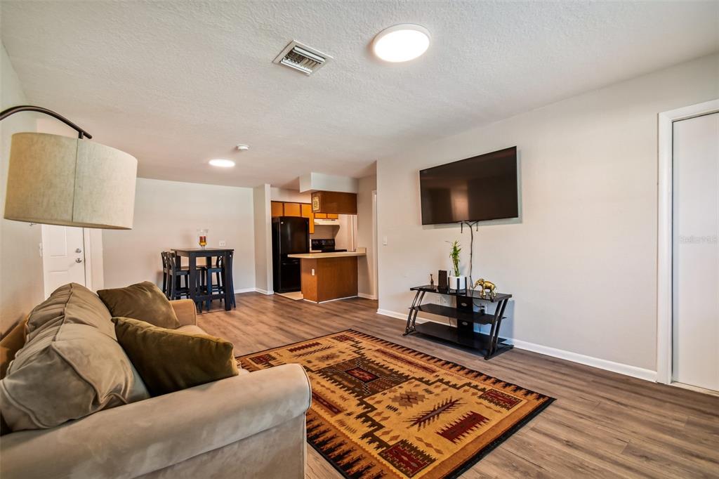Living room with a beige sectional, patterned rug, wall-mounted television, and open dining and kitchen areas.