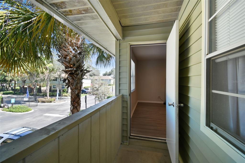 Covered balcony with green siding and an open door, overlooking palm trees and neighboring buildings.