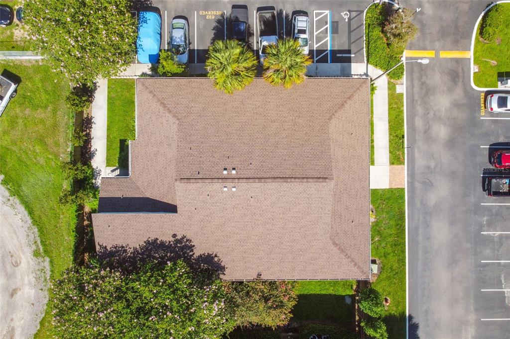 Aerial view of a residential building’s brown shingle roof beside parking spaces and landscaped grounds.