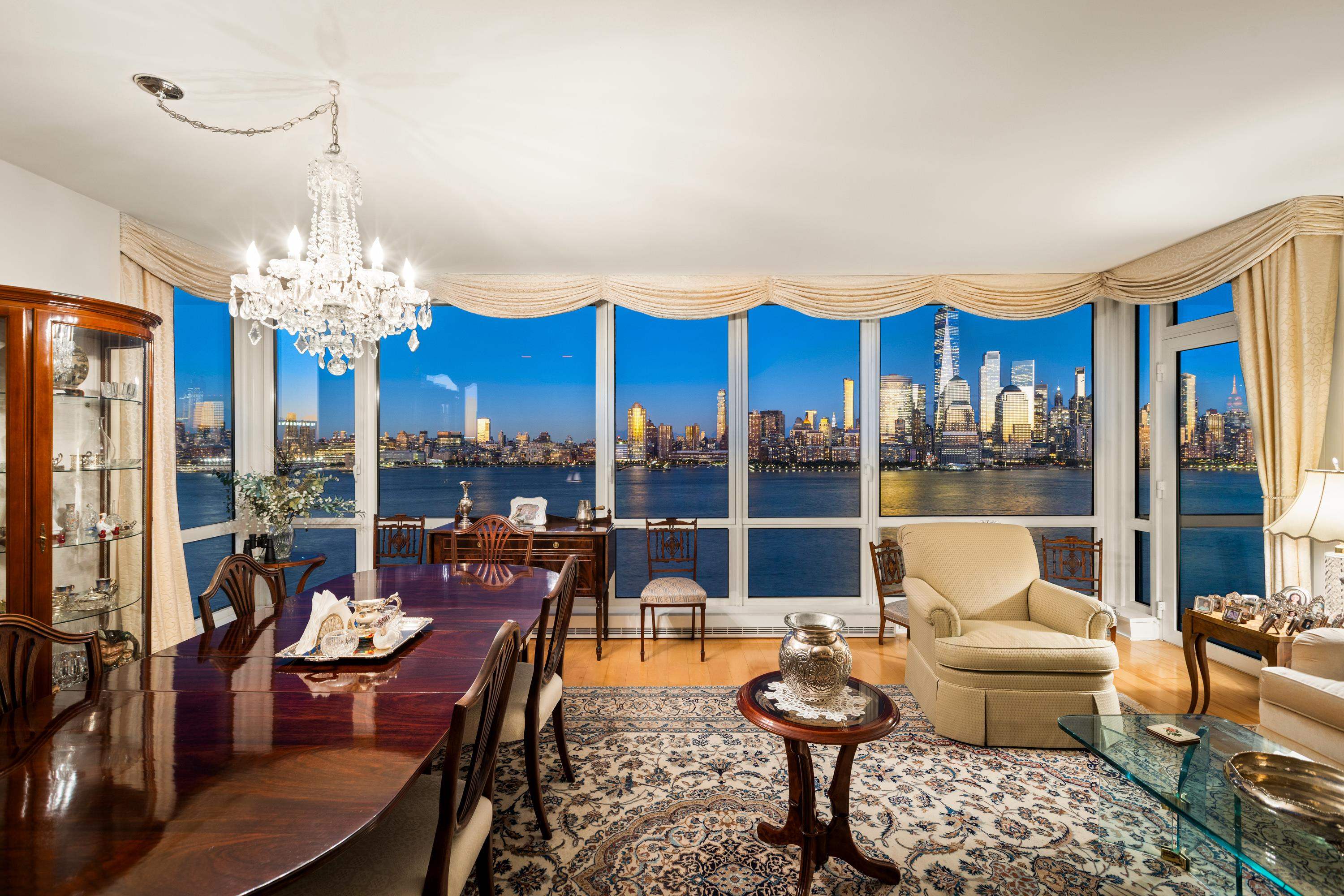 Furnished condo living and dining room with panoramic Hudson River and Manhattan skyline views at dusk.