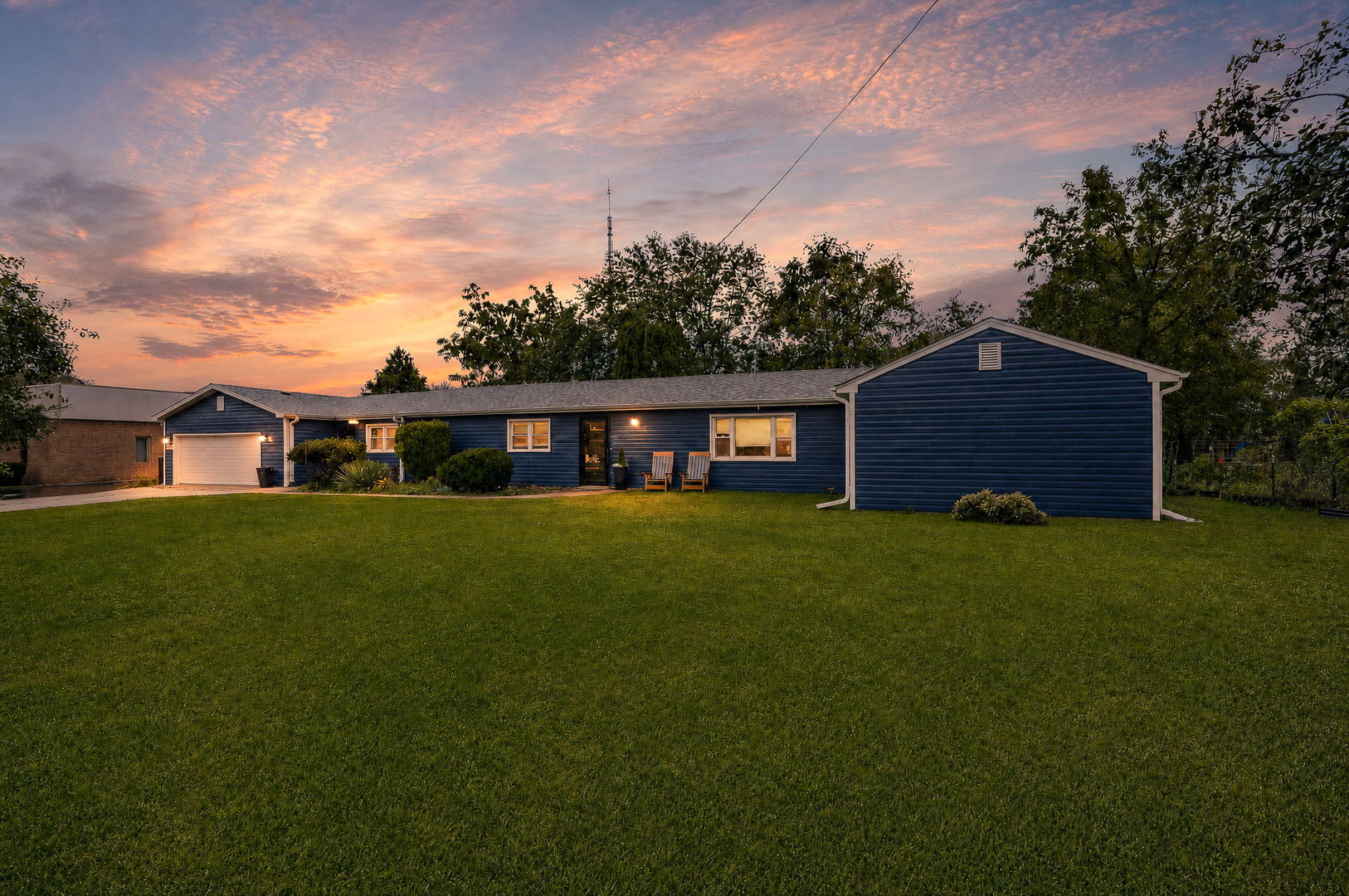 Blue single-story house with a white garage and broad lawn beneath a pink-orange sunset sky.