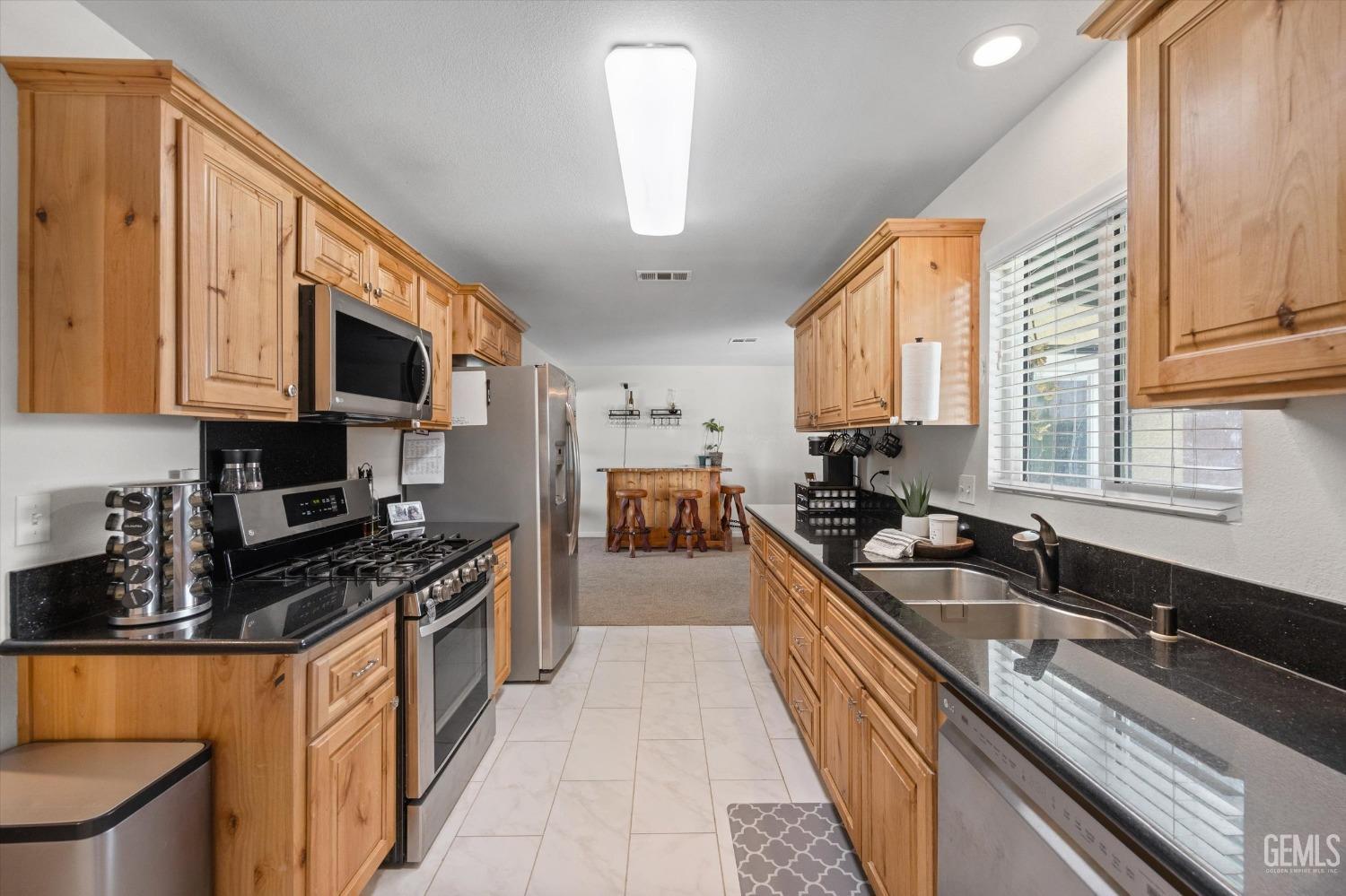Galley kitchen with honey-colored cabinets, black countertops, stainless steel appliances, and a sink beneath a window.