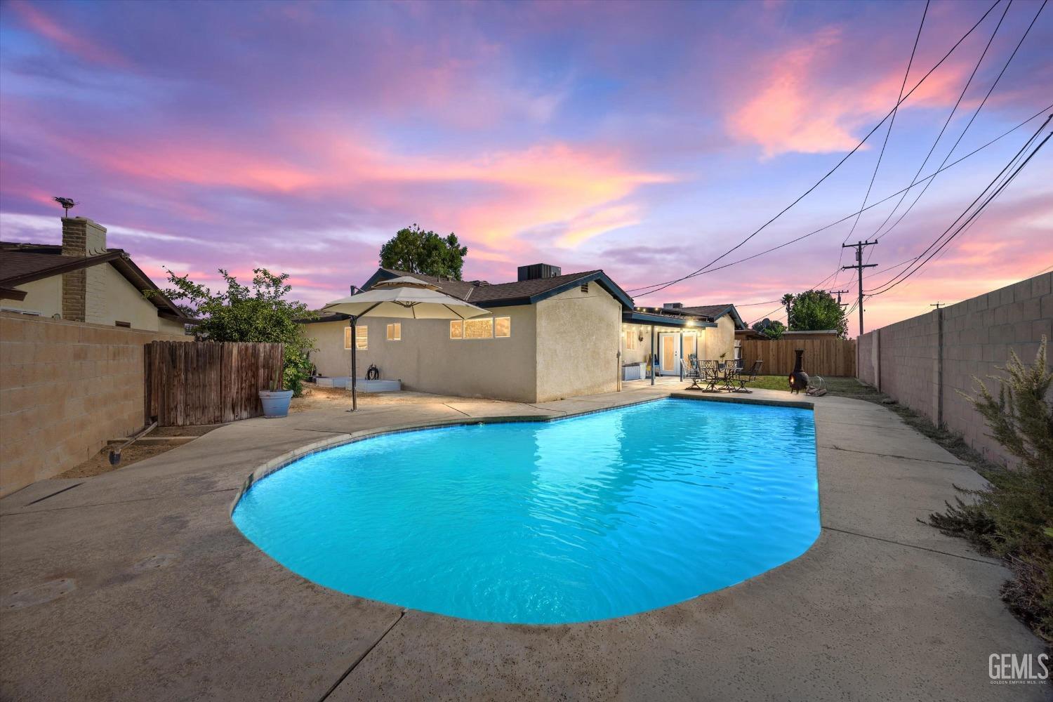 Backyard pool and concrete patio with umbrella and outdoor dining set at dusk.